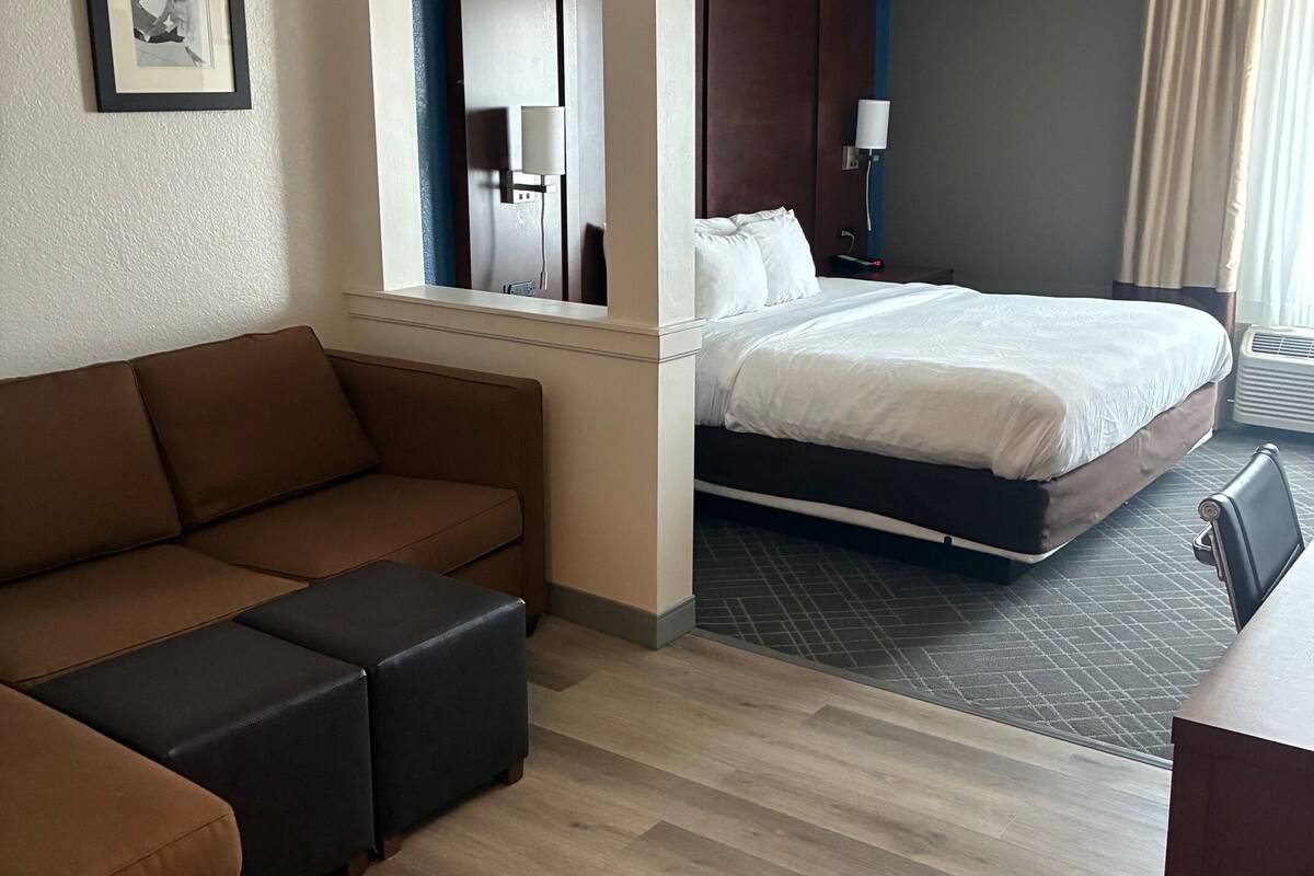 A hotel room with a brown sofa, a bed with white bedding, a mirror, and a nightstand with a lamp.