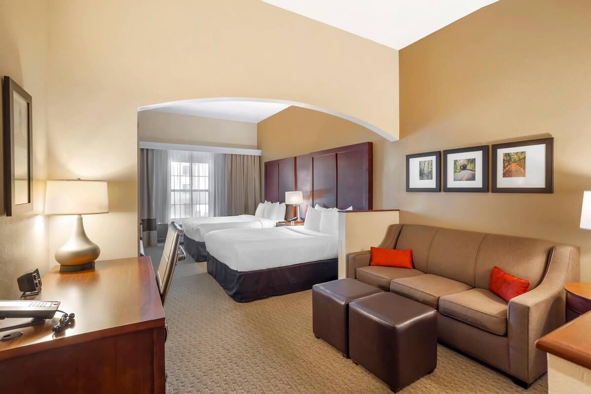 A hotel room with a bed, a sofa, a desk with a lamp, and a view of another room.