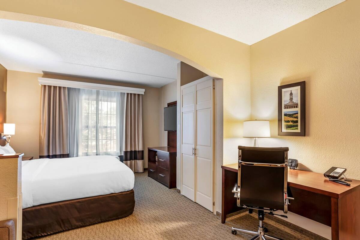 A hotel room with a bed, desk, chair, wardrobe, TV, and a framed picture on the wall.