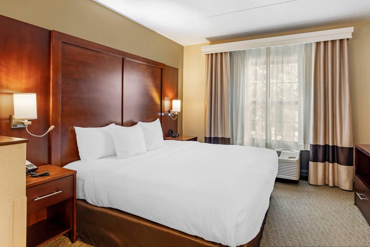 A hotel room with a large bed, a wooden headboard, a bedside table with a lamp, and a window with curtains.