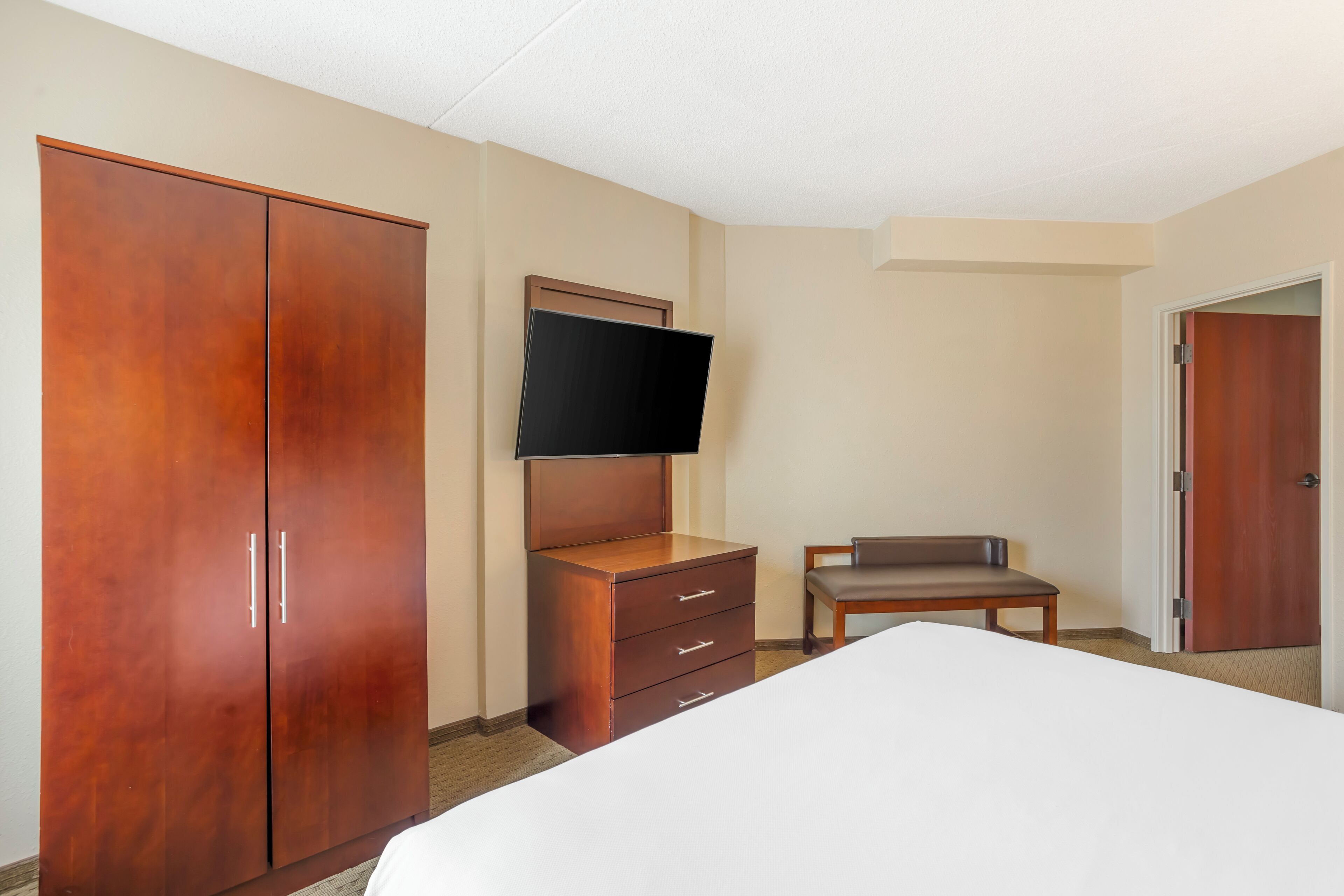 Habitación de hotel con una cama grande, un armario de madera, un televisor montado en la pared, una mesita y una puerta.