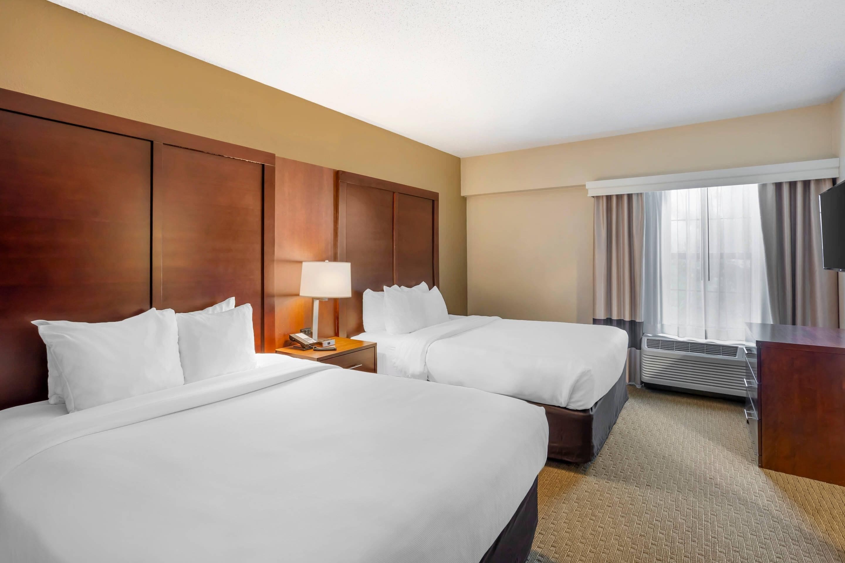A hotel room with two beds, a desk, a lamp, an air conditioning unit, and a television.