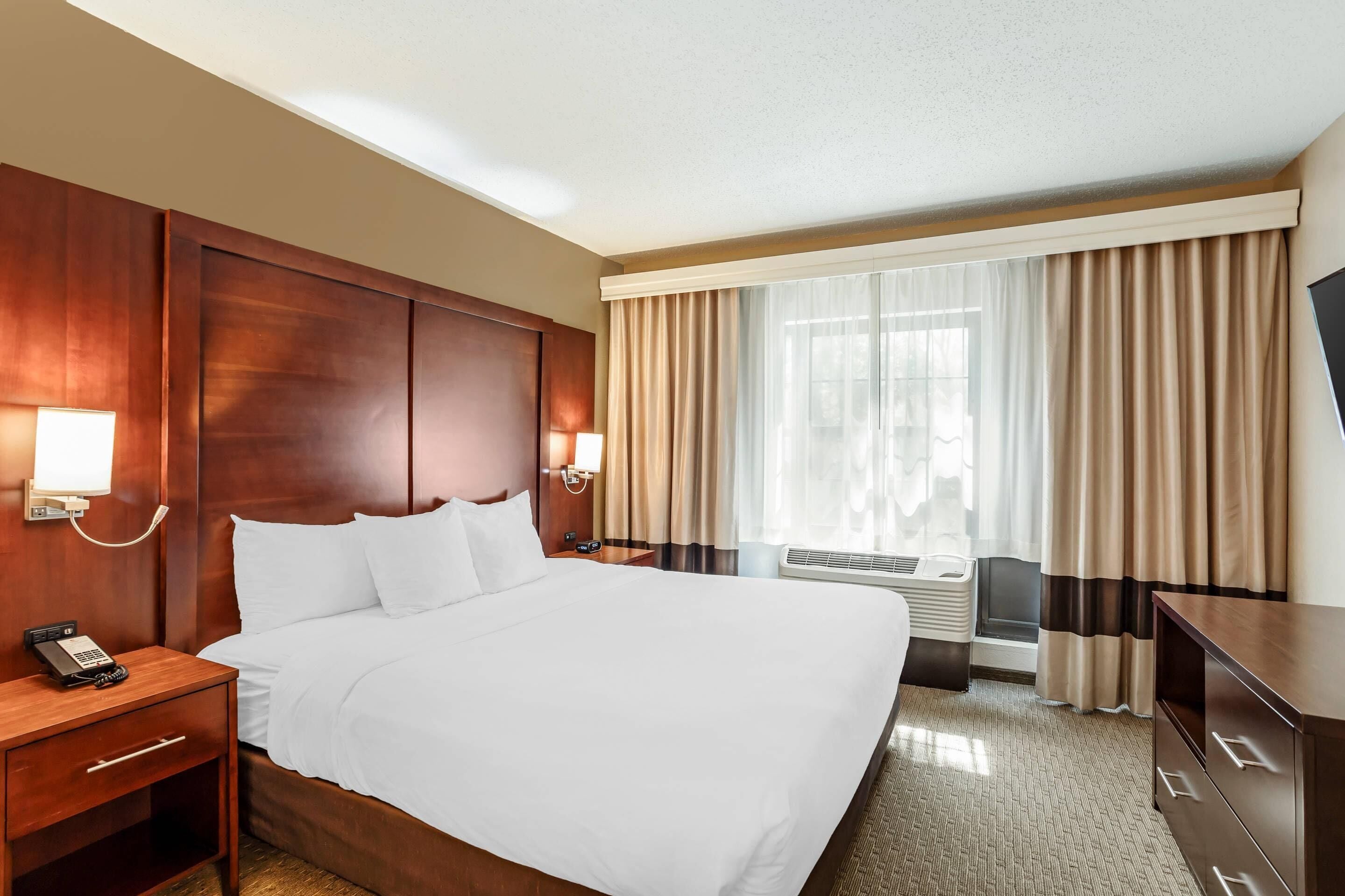 A hotel room with a large bed, wooden headboard, bedside lamps, a desk with a phone, and a large window with curtains.