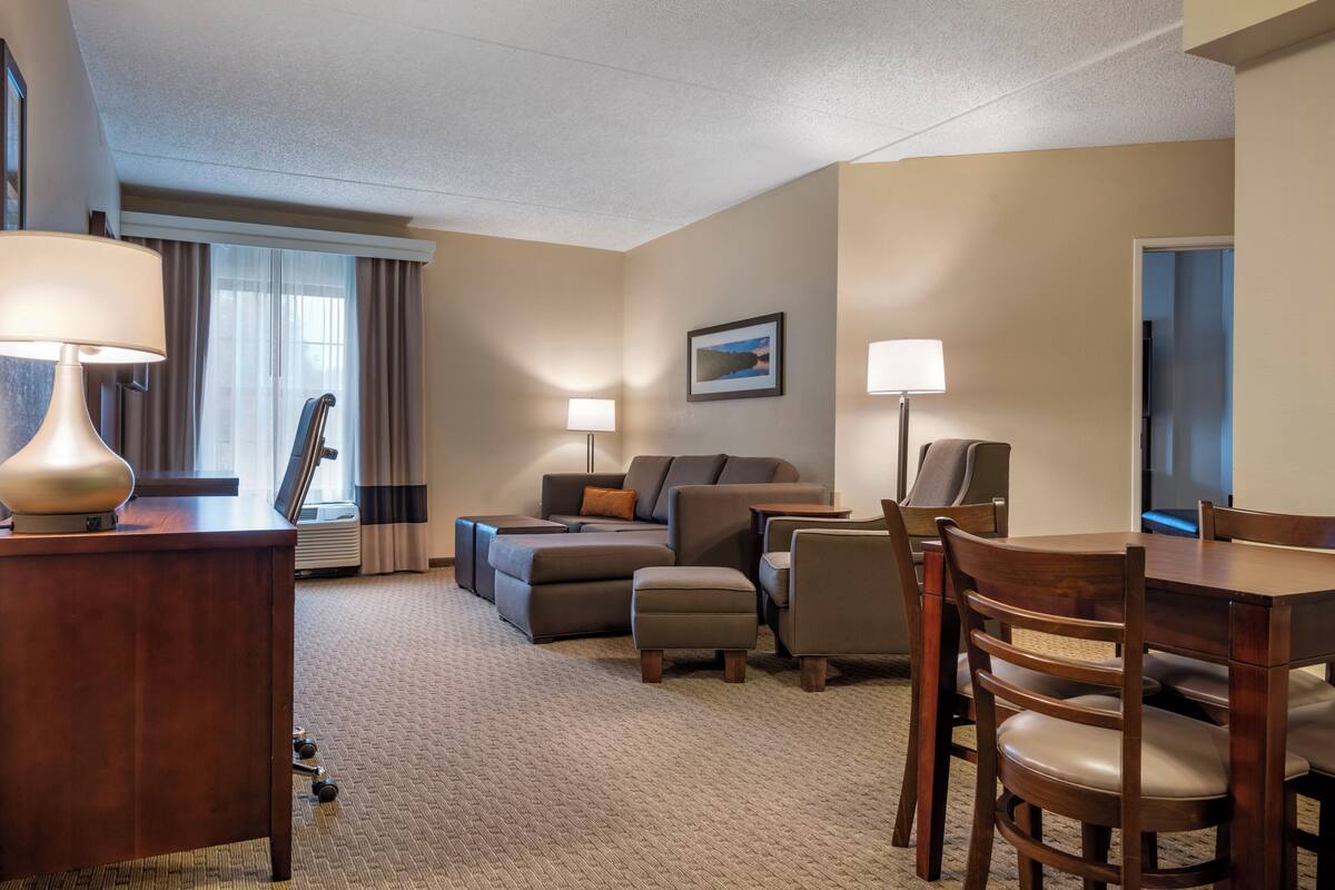A hotel room with a sofa, ottoman, dining table, chairs, lamps, and a picture on the wall.