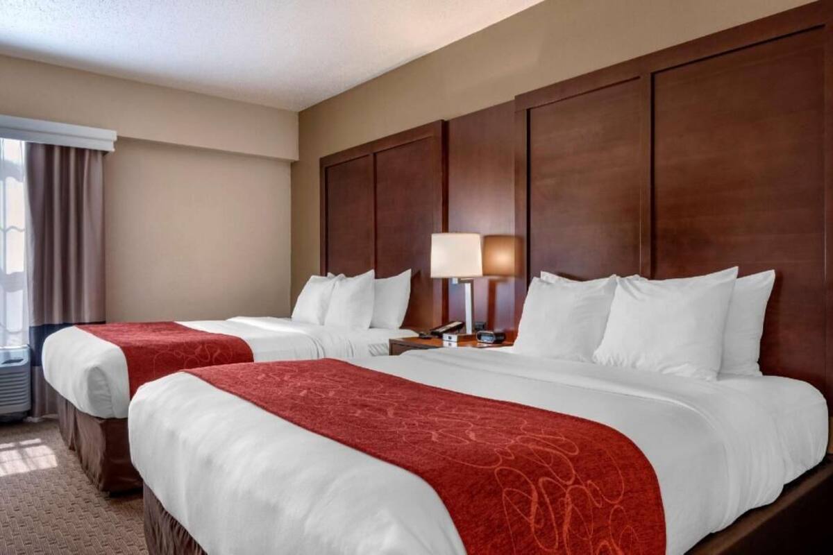 A hotel room with two beds, a large wooden headboard, a nightstand with a lamp, and a window with curtains.