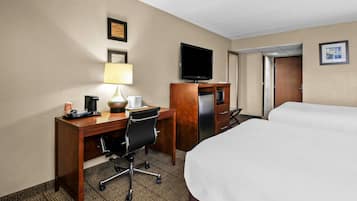 A hotel room with a bed, a desk with a chair, a television, a refrigerator, and a lamp.