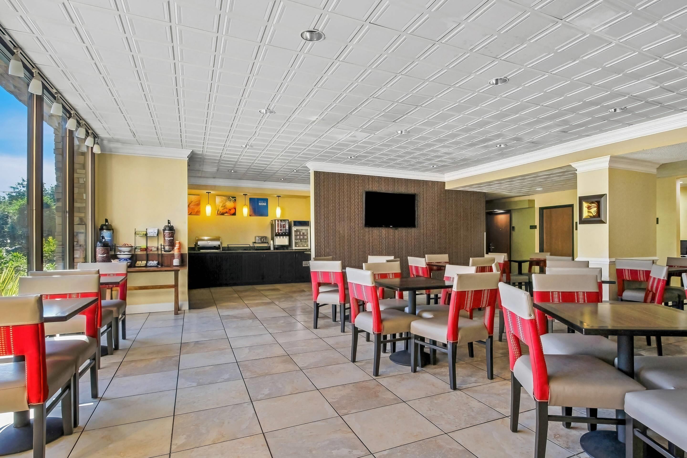 Un restaurant avec un plafond haut, un sol carrelé et des chaises recouvertes d’un tissu rouge et blanc. On y trouve un comptoir avec un menu et un téléviseur fixé au mur.