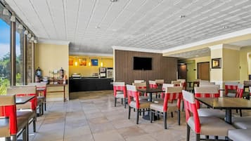 Un restaurant avec un plafond haut, un sol carrelé et des chaises recouvertes d’un tissu rouge et blanc. On y trouve un comptoir avec un menu et un téléviseur fixé au mur.