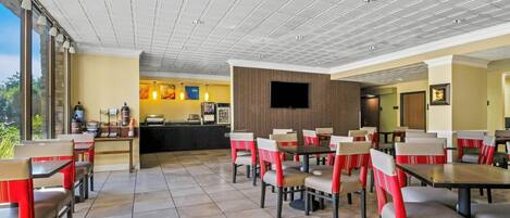 Un restaurante con techo alto, suelo de baldosas y sillas tapizadas en rojo y blanco. Se observa una barra con un menú y un televisor fijado a la pared.