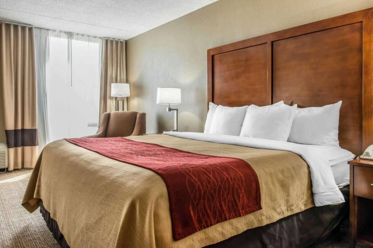 A hotel room with a large bed, a wooden headboard, two bedside lamps, a chair, and a view of the outdoors.