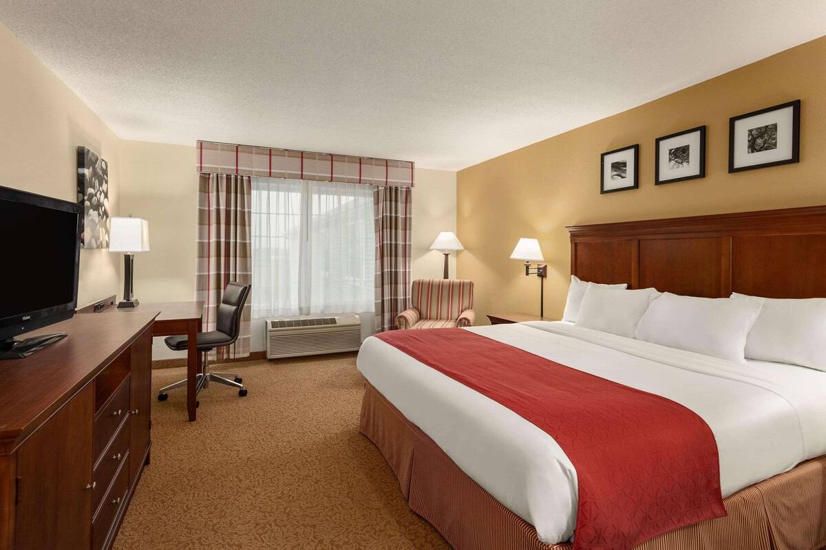 A hotel room with a bed, a desk with a TV, a chair, a window with curtains, and three framed pictures on the wall.