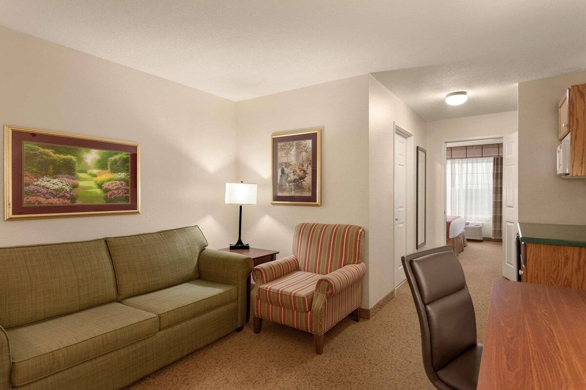 A hotel room with a green sofa, a striped armchair, a wooden dining table, and two framed pictures on the wall.
