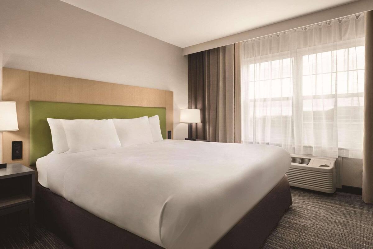 A hotel room with a large bed, a green headboard, two bedside tables with lamps, a window with curtains, and a wall-mounted air conditioning unit.