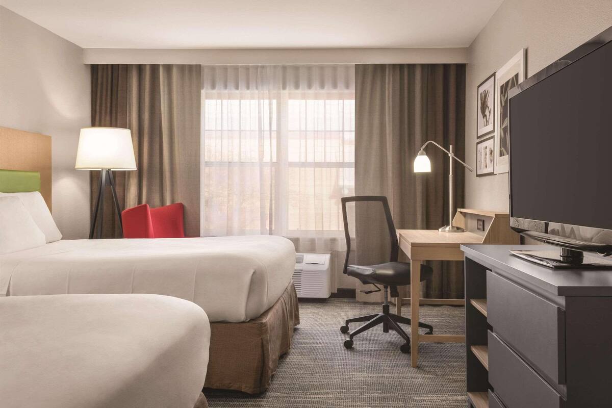 A hotel room with two beds, a desk, a chair, a TV, and a lamp.