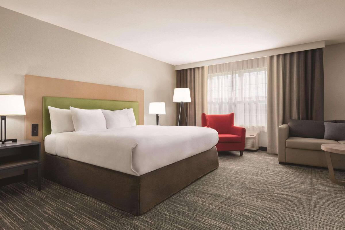 A hotel room with a large bed, a red armchair, a grey sofa, and a lamp.