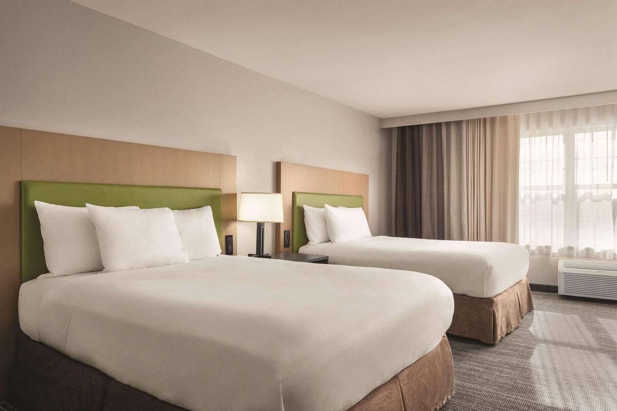 A hotel room with two beds, a large window with curtains, and an air conditioning unit.
