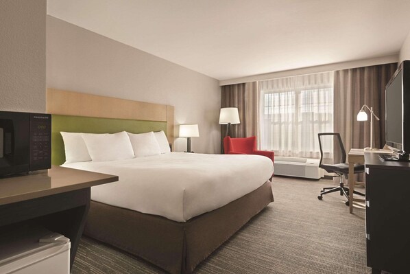 Quarto de hotel com cama, escrivaninha, cadeira, televisão, luminária e janela com cortinas.
