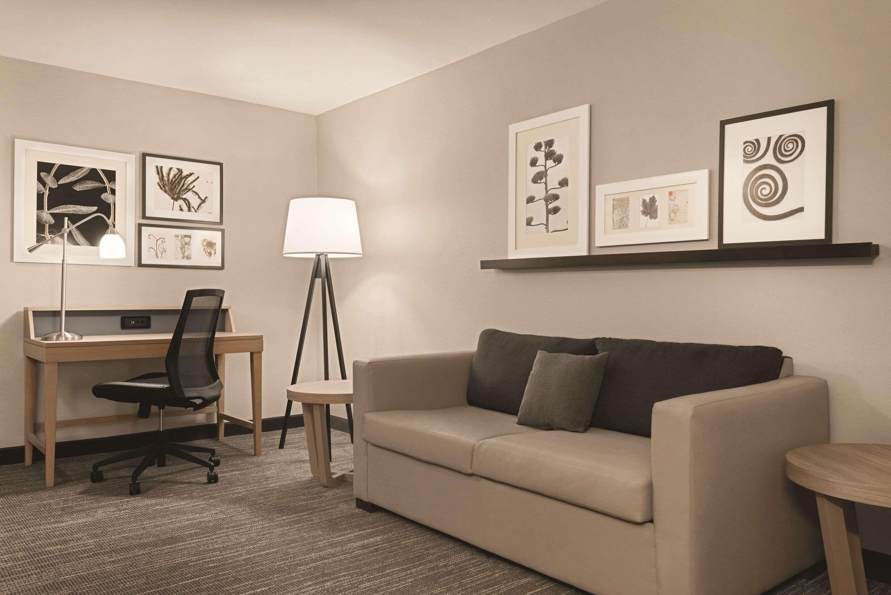 A hotel room with a desk, a chair, a sofa, a lamp, and framed artwork on the walls.