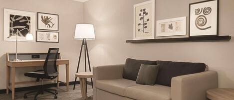 A hotel room with a desk, a chair, a sofa, a lamp, and framed artwork on the walls.