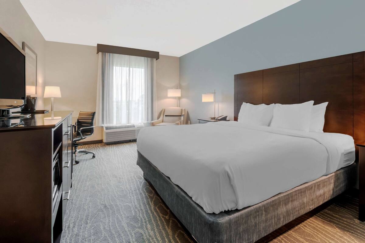 A hotel room with a large bed, a desk with a computer, two chairs, a TV, and a window with curtains.