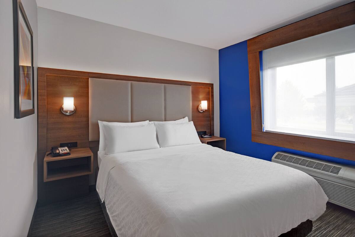 A hotel room with a large bed, two bedside tables, a nightstand, a wall-mounted lamp, and a window with blinds.