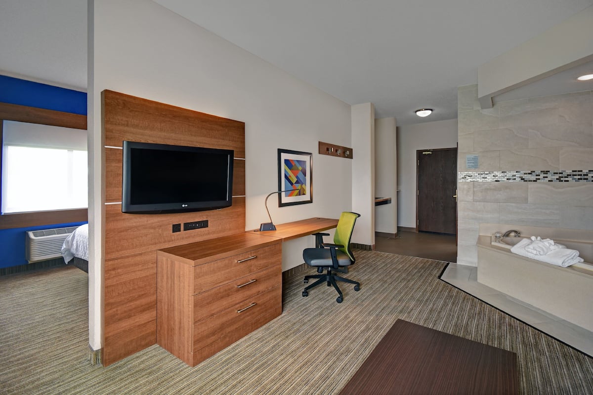 A modern hotel room with a wooden desk, a flat-screen TV, a green office chair, and a bathroom with a large mirror and a bathtub.