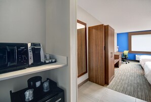 Quarto de hotel com cozinha compacta, uma cama, escrivaninha com cadeira e janela.