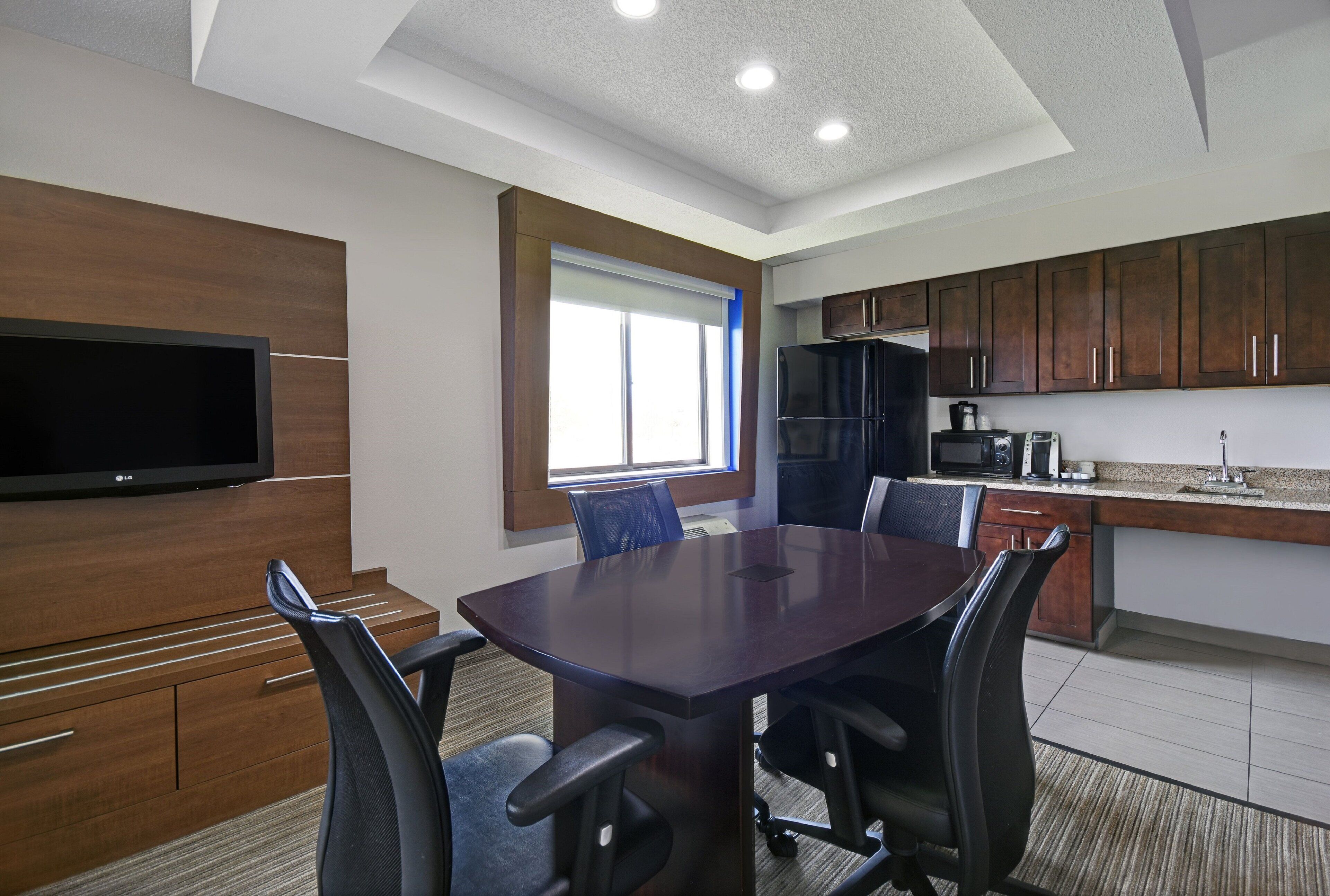 A conference room with a dark wooden table, black office chairs, a flat-screen TV, and a kitchen area with wooden cabinets and appliances.