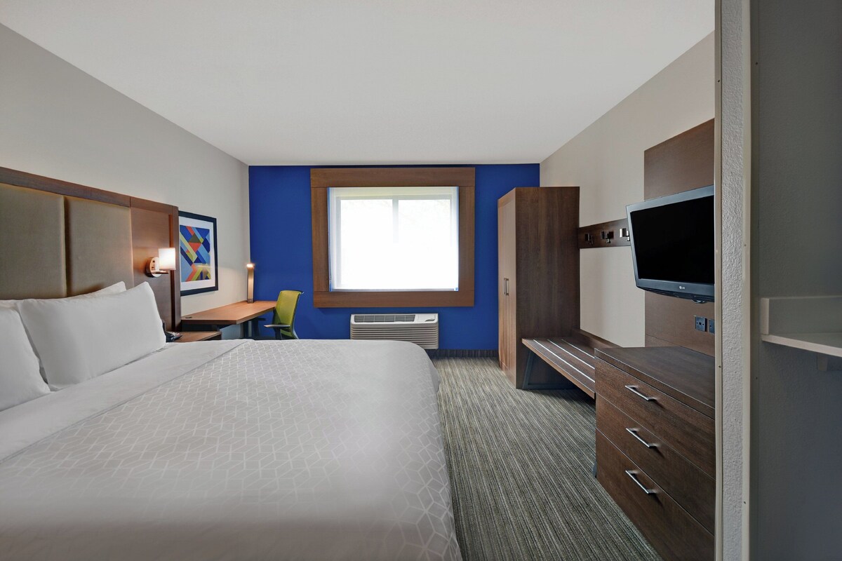 A hotel room with a large bed, a desk with a chair, a television, and a window with blinds.