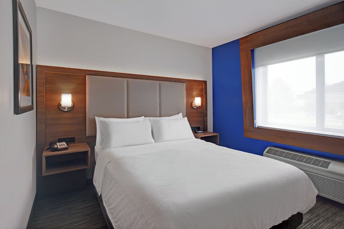 A hotel room with a large bed, two bedside tables, a nightstand, a wall-mounted lamp, and a window with blinds.