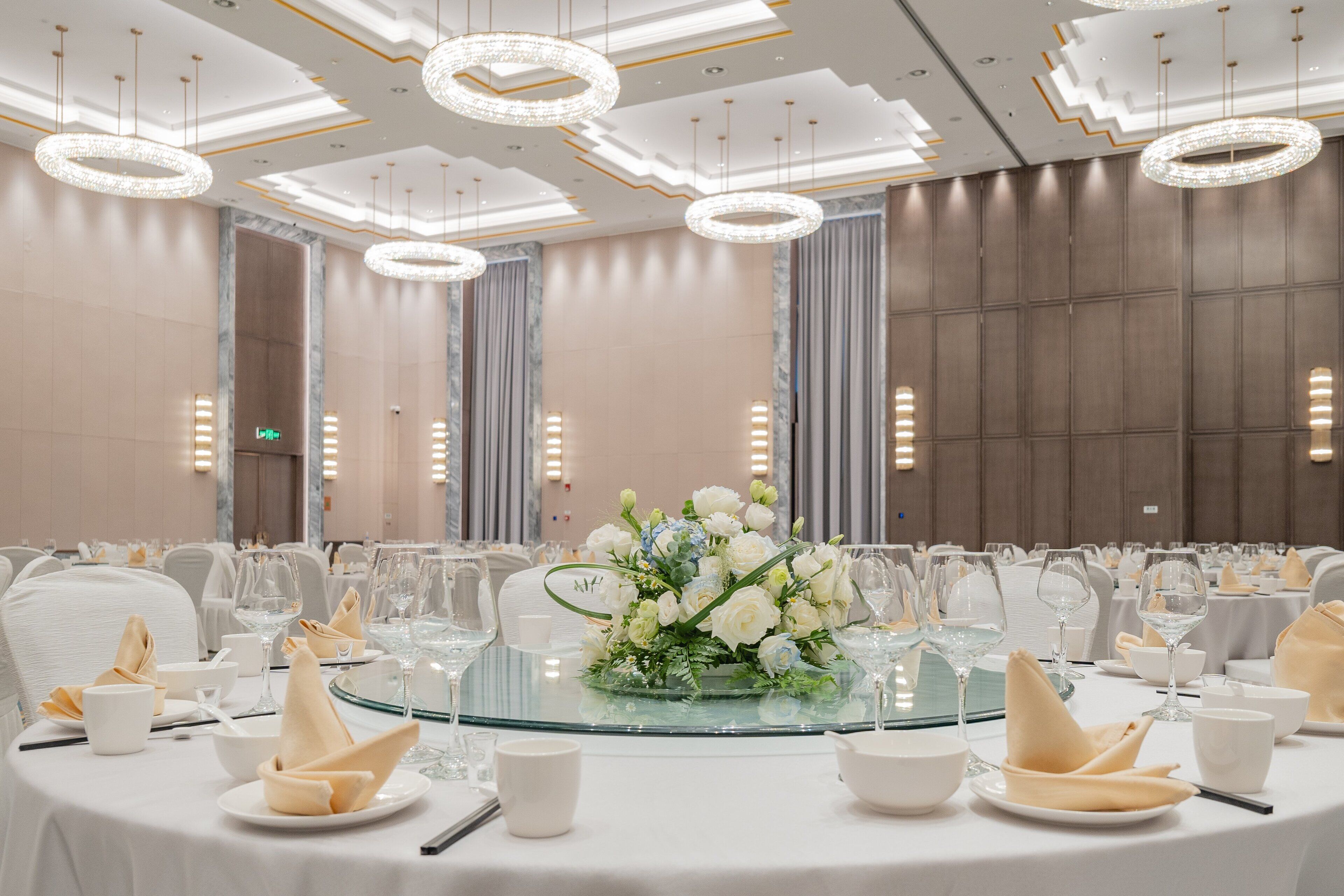 Un salón de banquetes con mesas redondas dispuestas para un evento formal, con tapas de cristal, manteles blancos y centros de mesa florales.