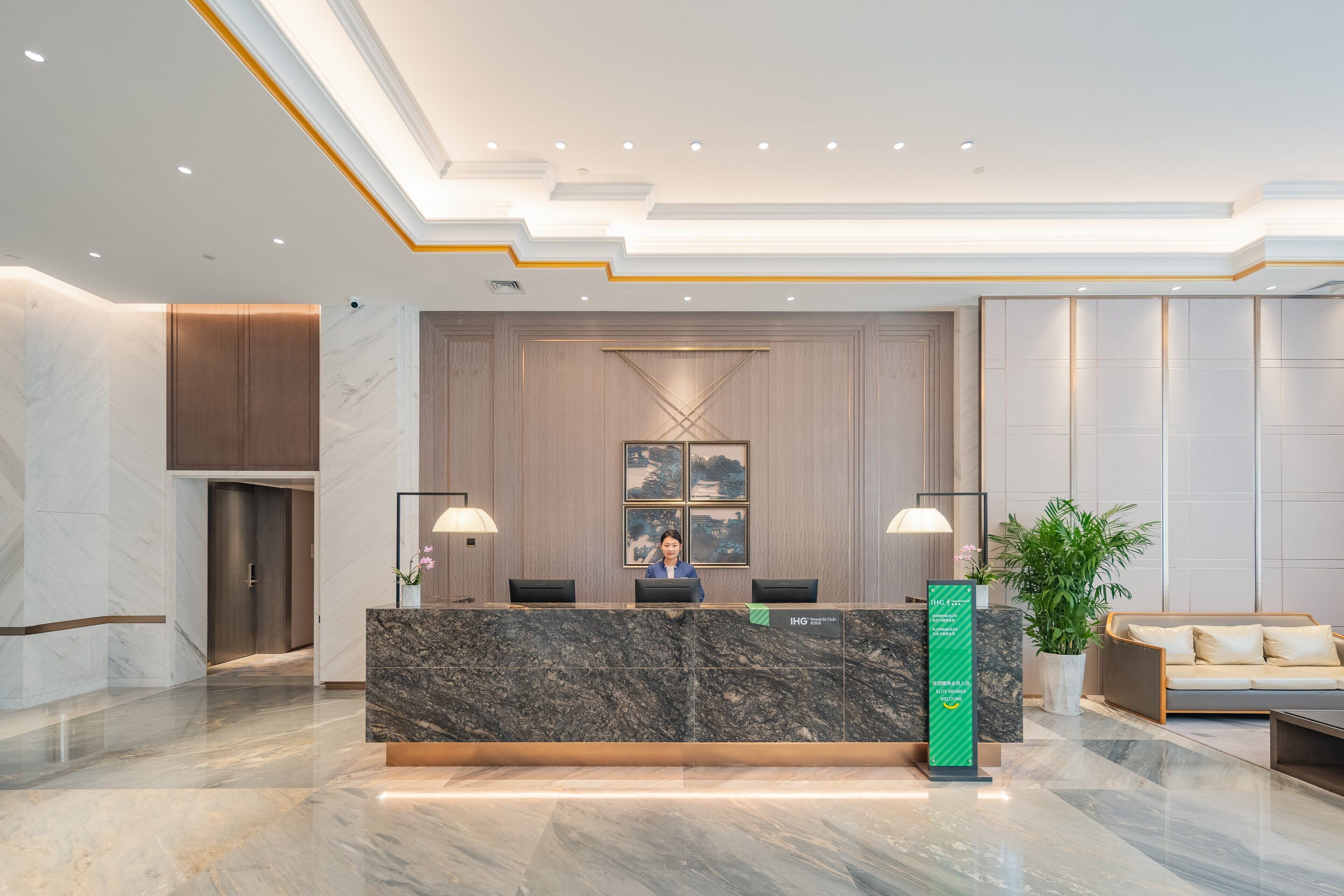 A hotel reception area with a marble reception desk, two black chairs, a reception counter, a large potted plant, and a sofa with cushions.