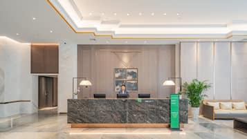 A hotel reception area with a marble reception desk, two black chairs, a reception counter, a large potted plant, and a sofa with cushions.