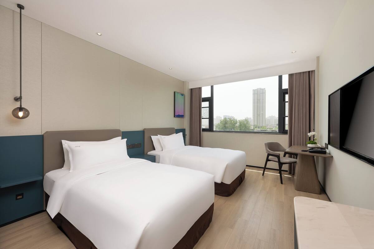 A hotel room with two beds, a desk, a TV, and a window with a city view.