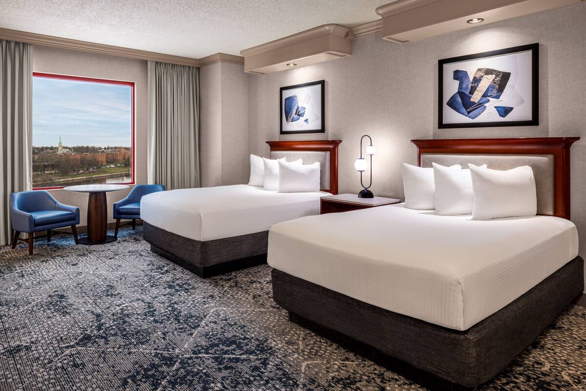 A hotel room with two beds, a view of a cityscape, a dining table, and blue chairs.