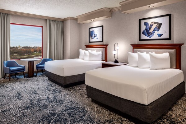 A hotel room with two beds, a view of a cityscape, a dining table, and blue chairs.