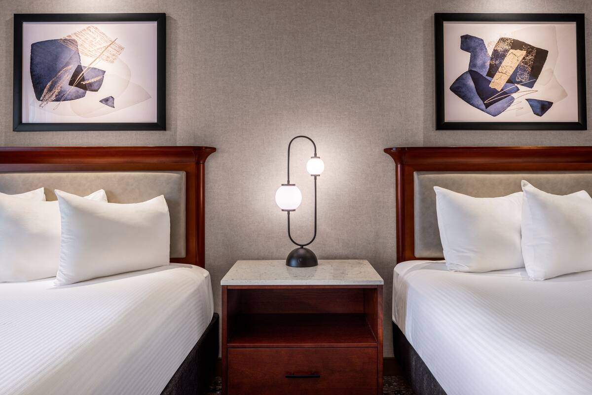 A hotel room with two beds, a nightstand with a lamp, and artwork on the walls.
