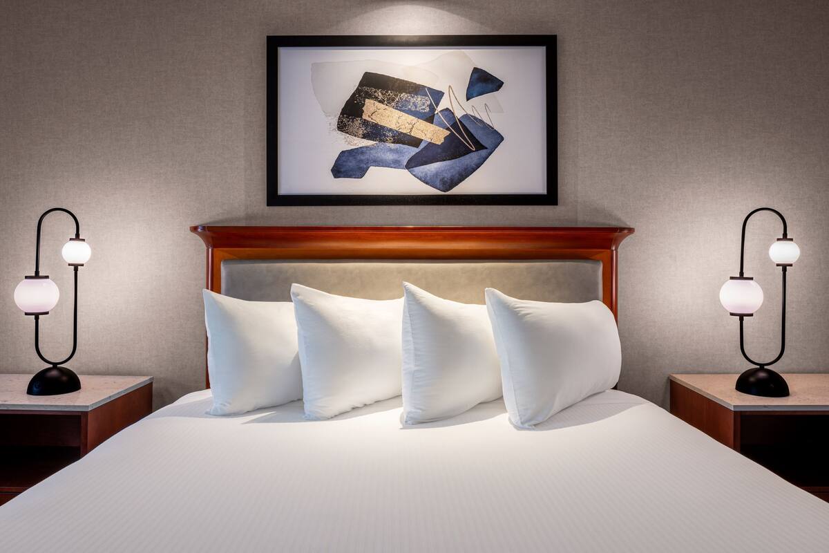 A neatly made bed with white pillows and a framed abstract artwork above it. There are two bedside tables with lamps on either side of the bed.