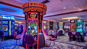 Un salón de casino con diversas máquinas tragamonedas, incluyendo los juegos 'DOLLAR STORM' y 'JACKPOT', y múltiples mesas de juego.