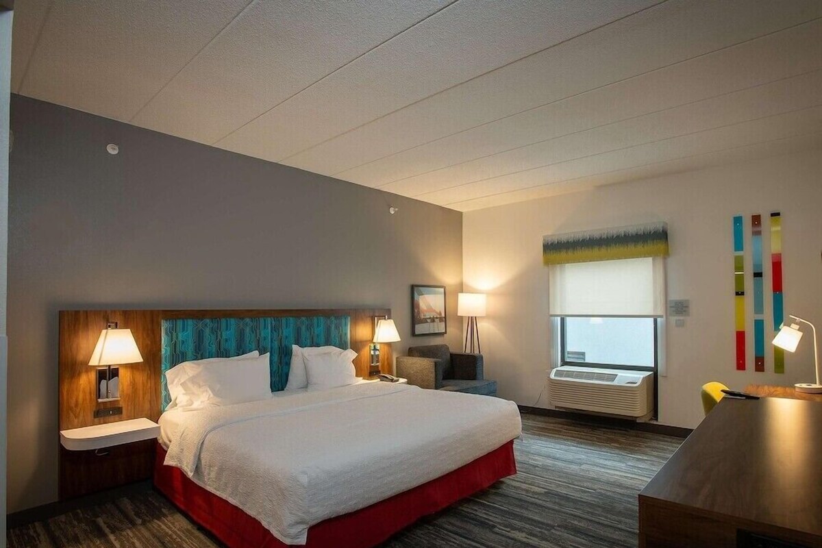 Habitación de hotel con una cama grande, dos mesitas de noche con lámparas, un escritorio, una silla, un ventanal con persianas y una obra de arte abstracta y colorida en la pared.