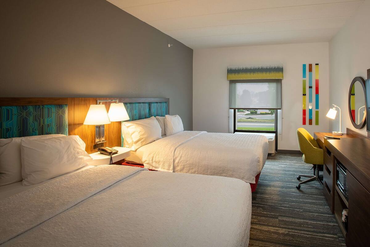 Quarto de hotel com duas camas, uma escrivaninha com cadeira, uma janela com vista e decoração colorida nas paredes.