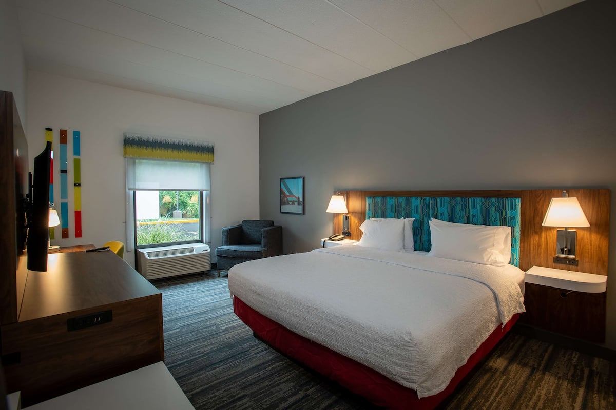 Quarto de hotel com uma cama grande, duas mesas de cabeceira com luminárias, uma escrivaninha, uma cadeira, uma janela com persianas e um ar-condicionado fixado na parede.