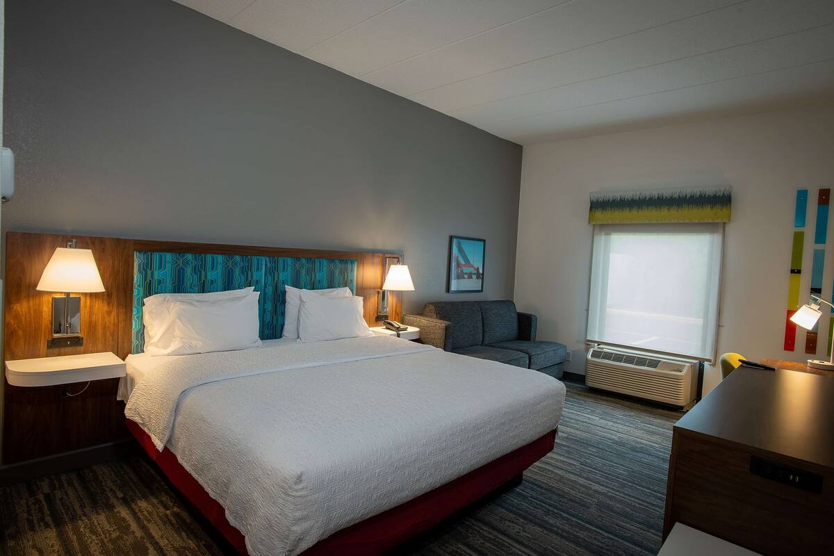 Quarto de hotel com uma cama grande, duas mesas de cabeceira com abajures, um sofá cinza, uma escrivaninha com abajur e uma janela com persianas.