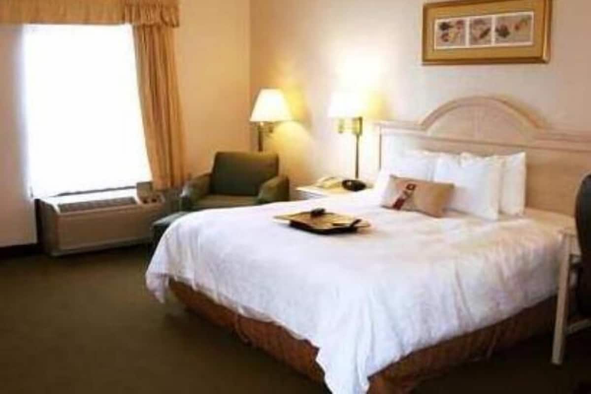 Quarto de hotel com cama, uma cadeira, escrivaninha e janela com cortinas.