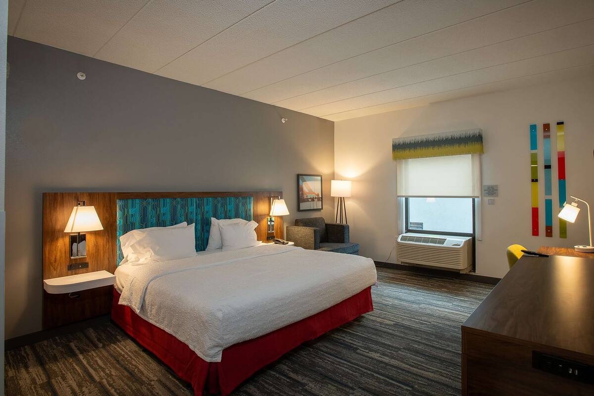 Quarto de hotel com uma cama grande, duas mesas de cabeceira com abajures, uma escrivaninha, uma cadeira, uma janela com vista e uma parede com decoração colorida.