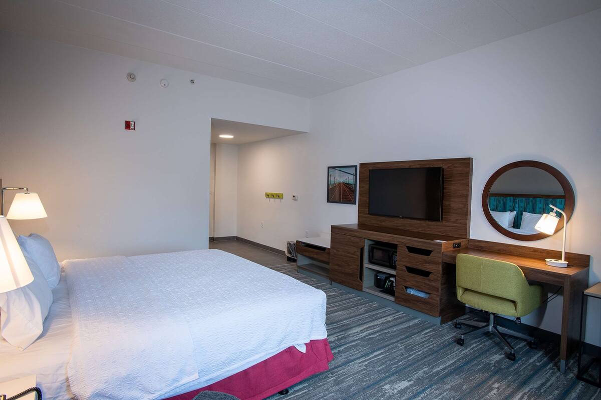 Quarto de hotel com cama, escrivaninha, cadeira, televisão e luminária.