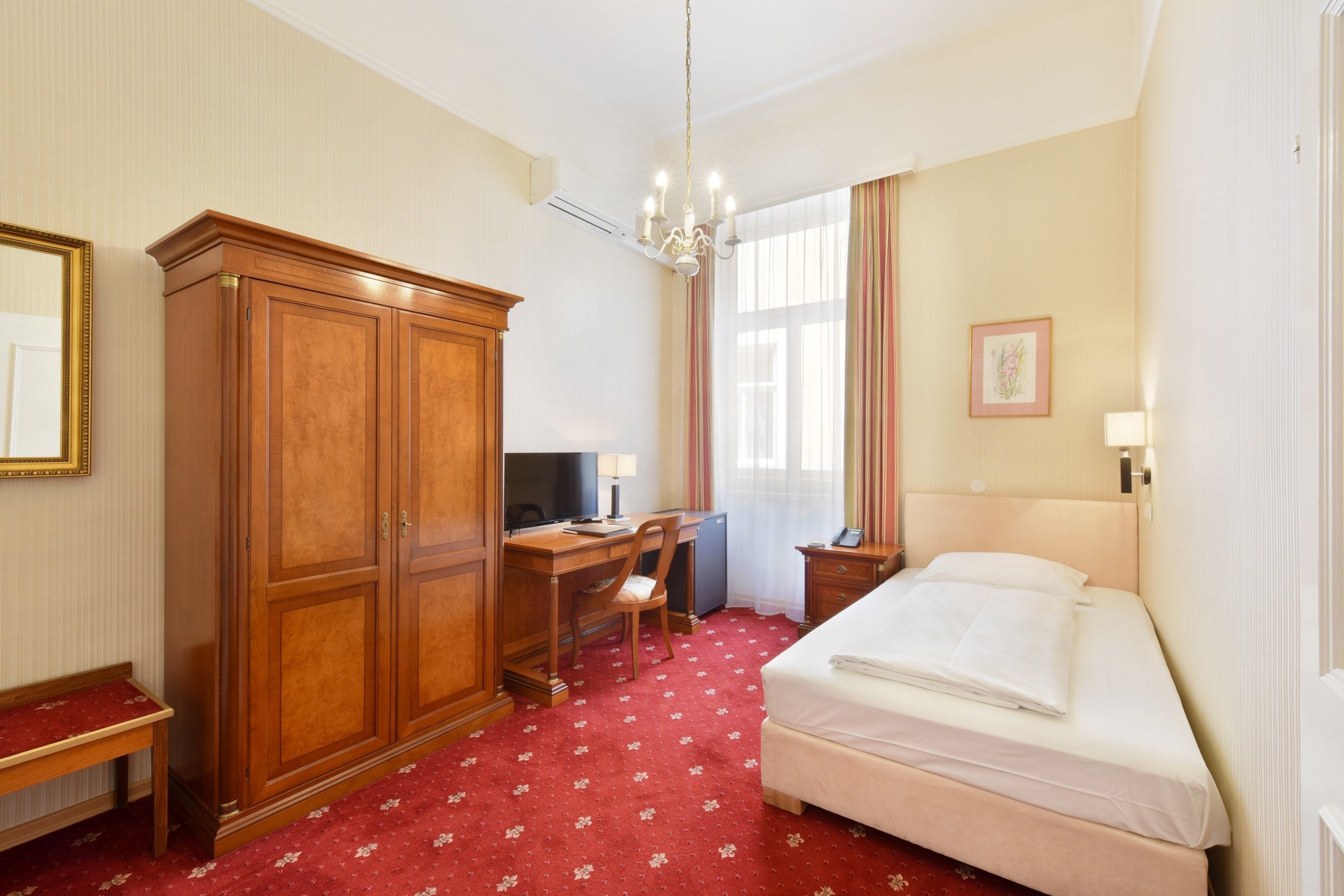 Ein Hotelzimmer mit Bett, Schreibtisch mit Stuhl, Kleiderschrank, Fernseher und einem Fenster mit Vorhängen.