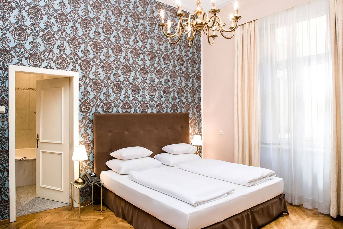 Une chambre d’hôtel avec un grand lit, un lustre, un papier peint à motifs et une vue sur l’extérieur à travers de fines rideaux.