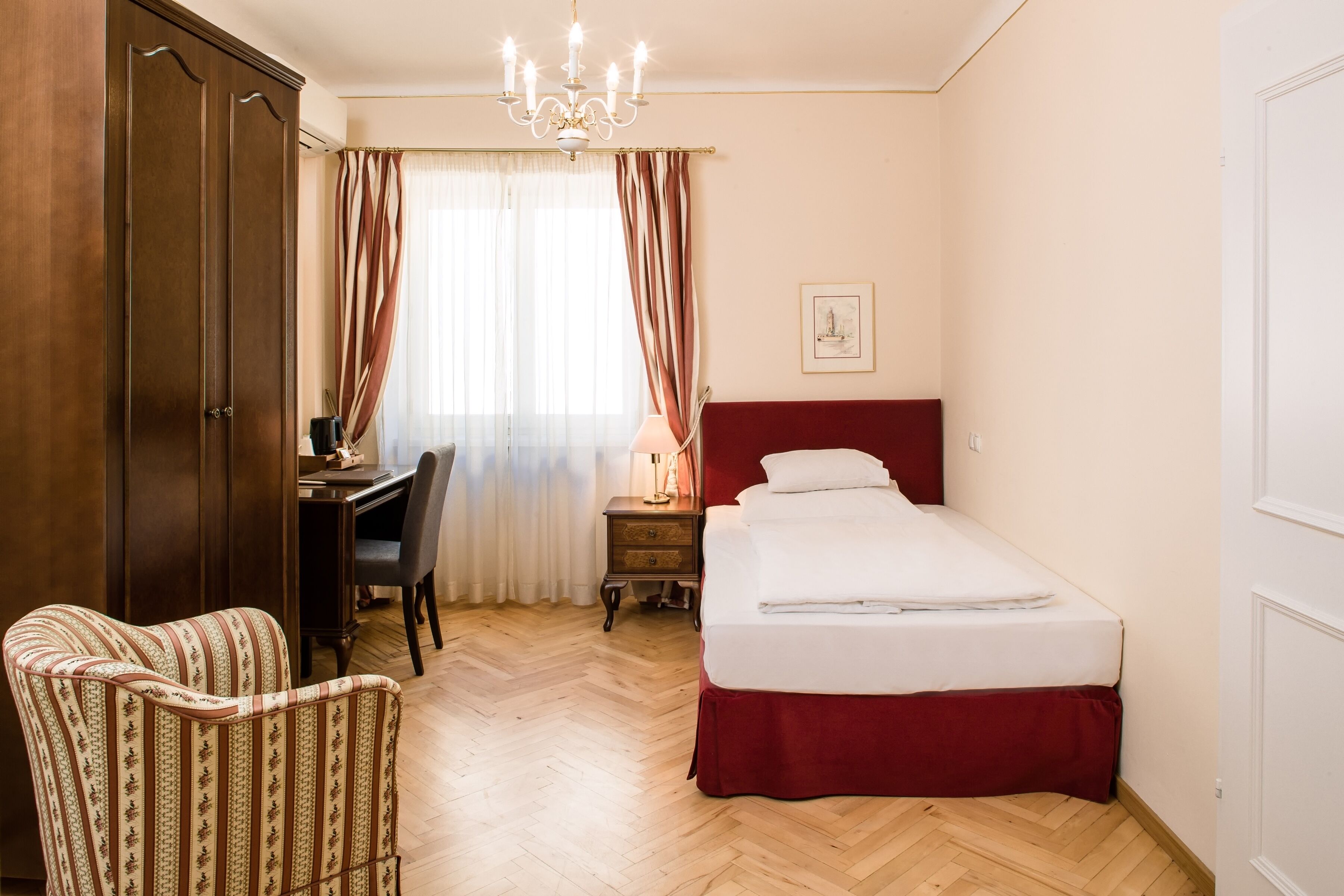 A hotel room with a wooden wardrobe, a desk with a chair, a bed with a red headboard, and a chandelier.