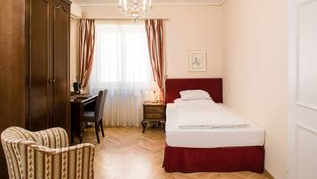 A hotel room with a wooden wardrobe, a desk with a chair, a bed with a red headboard, and a chandelier.